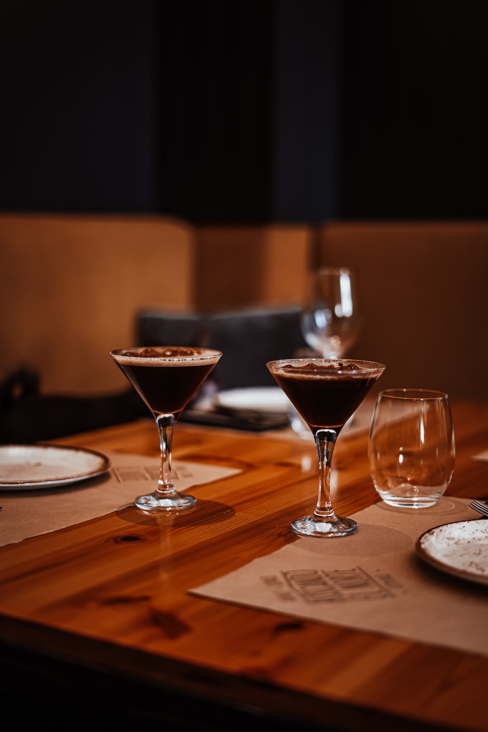 Dos cócteles espresso martini sobre la mesa de madera, con el mantel de BEN y la banqueta en penumbra al fondo